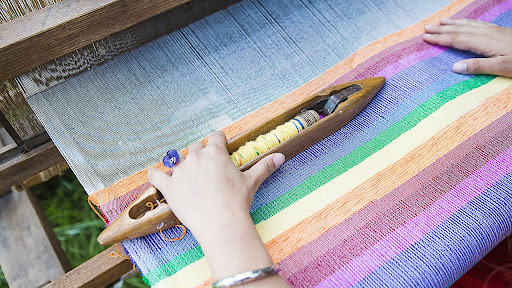 Person with a purple ring hand weaving a rainbow-colored rug on a loom.