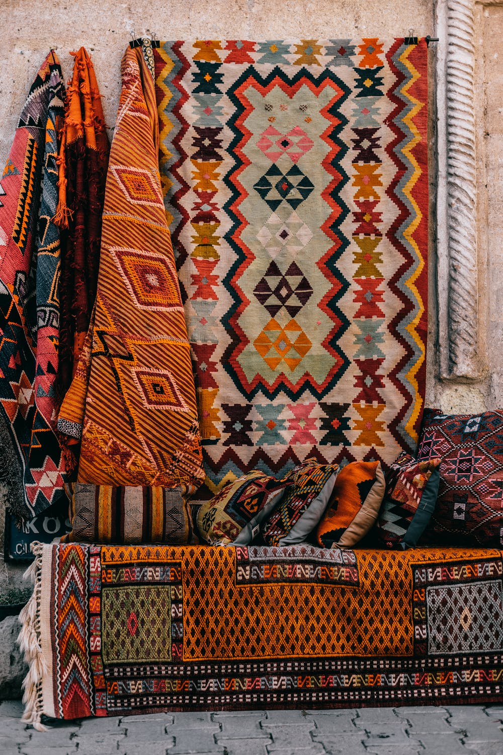 A multi-colored kilim is displayed in front of a stone wall surrounded by other rugs and tapestries at a market.